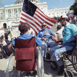 pictured are 3 people who use a wheelchair with an American flag that has a handicapped icon on it