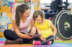 Photo of a young girl sitting on the floor reading a book with an adult. Wheelchair in the background.