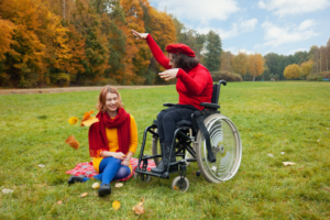 two woman outside during fall throwing leaves. One woman is seated in a wheelchair and the other is sitting on the grass.